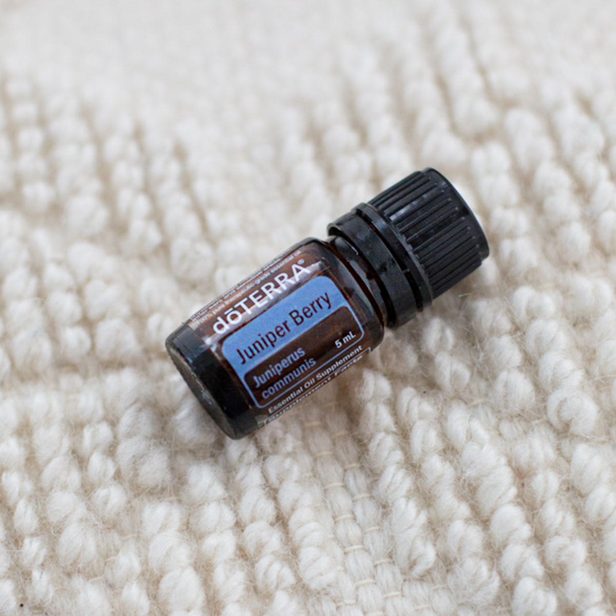 Juniper Berry essential oil bottle resting on a white patterned cloth. Wondering how to use Juniper Berry oil? Juniper oil can be used to tone the skin, to create a refreshing and grounded environment, or for internal benefits. 