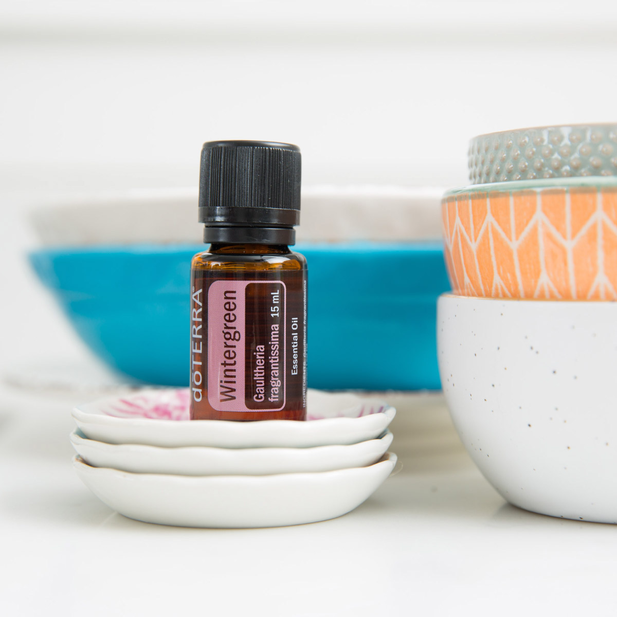 Bottle of Wintergreen oil sitting on a white dish, surrounded by other kitchen dishes. Wondering how to use Wintergreen essential oil? Try adding a few drops of Wintergreen oil to your bathwater for a soothing bath. 