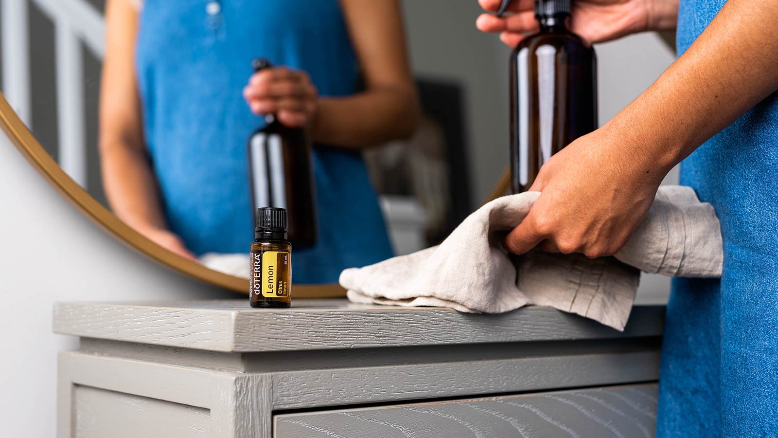 A woman holding a glass spray bottle and rag, about to spray a a gray table. doTERRA Lemon essential oil sitting on the side of the table. 