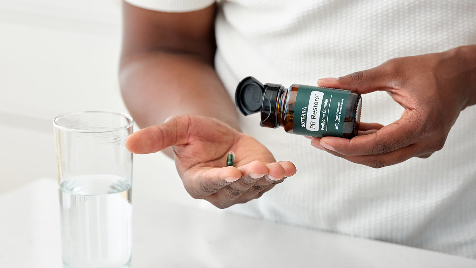 Man holding the PB Restore ProBiome Complex bottle, gently pouring the pills out. A clear glass of water sits on a white counter. 