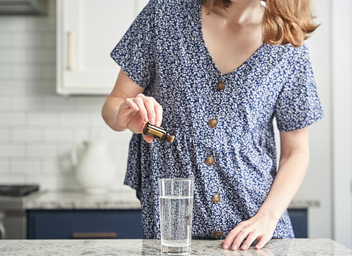A woman standing next to a marble kitchen counter, her hand rested on the counter, and her other hand holding a bottle of doTERRA Ginger essential oil, pouring it into a glass of water.  