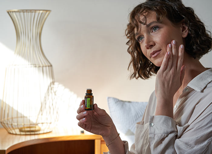 A woman sitting, holding doTERRA Tea Tree essential oil, and applying the oil to the side of her face.  