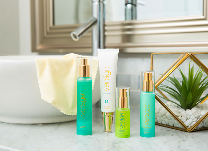 doTERRA Veráge Skin Care Collection sitting on a marble counter. Beside them is a green plant. Behind the collection is a large, bowl-shaped sink and a mirror.  