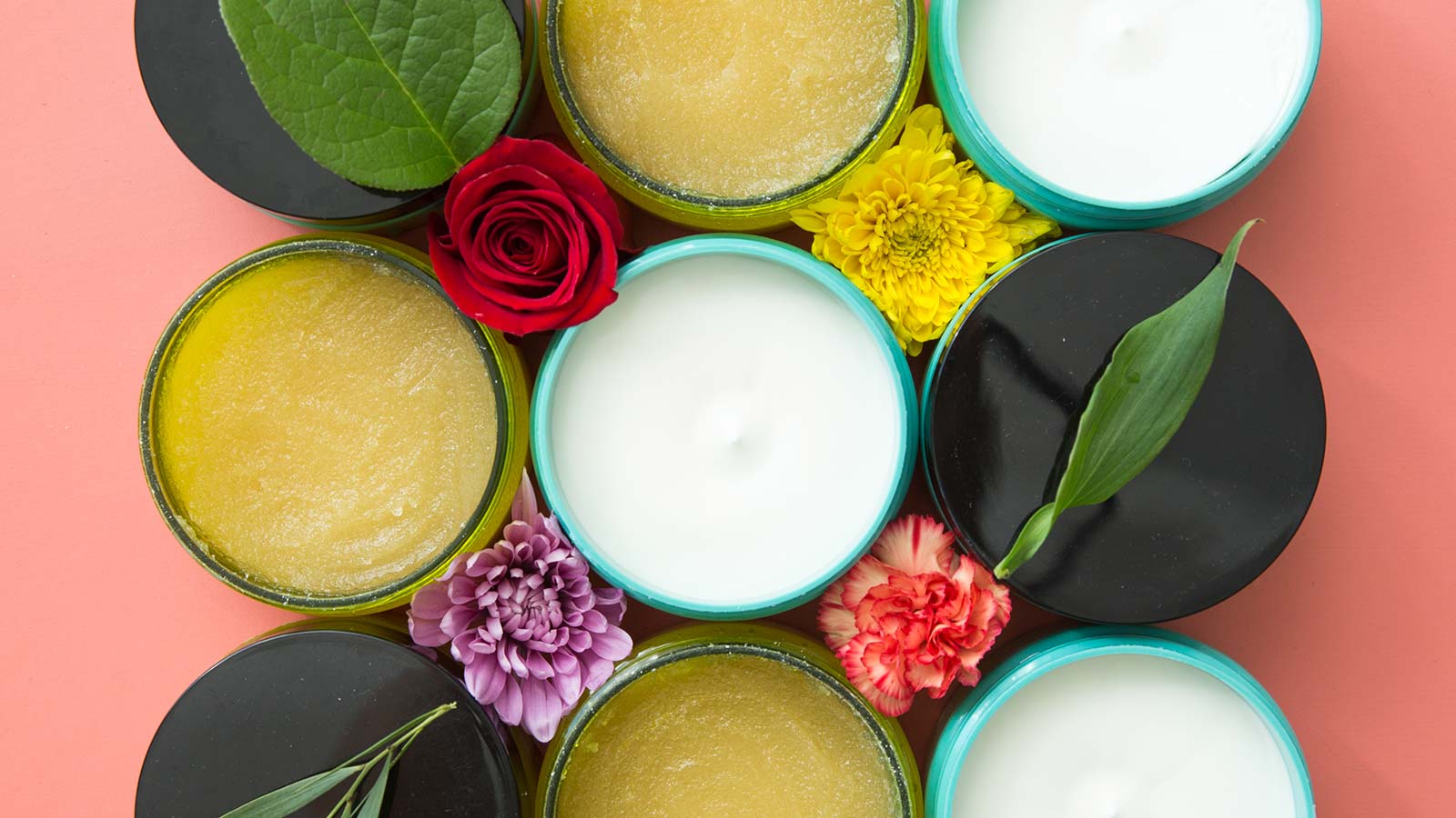 Overhead shot of nine bottles of lotion and body scrub with some lids open and some closed. The containers create a square with leaves and flowers between them.  