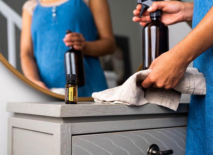 A woman holding a glass spray bottle and rag, about to spray a a gray table. doTERRA Lemon essential oil sitting on the side of the table. 