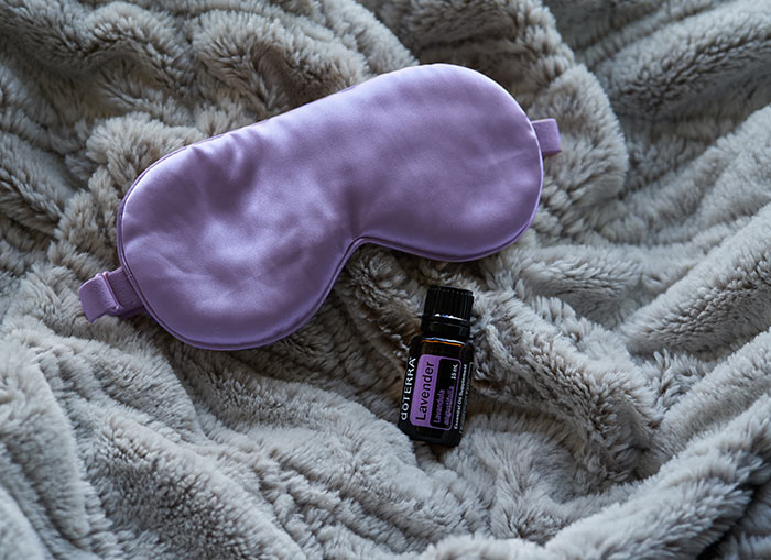 doTERRA Lavender essential oil lays on a fluffy, gray blanket, with a purple sleep mask above it.  
