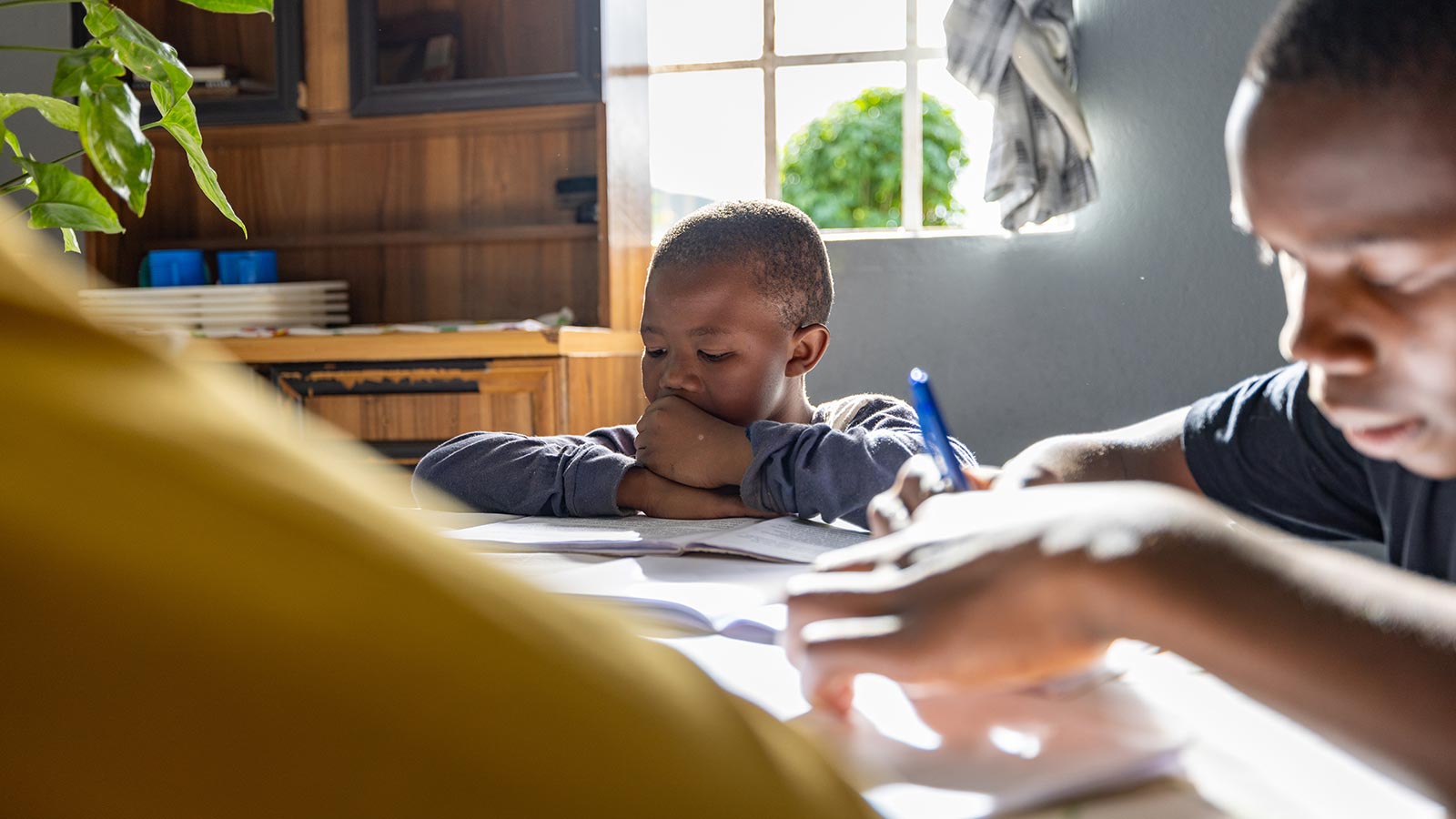 Two boys kneeling on the ground, up against a table, doing homework. 