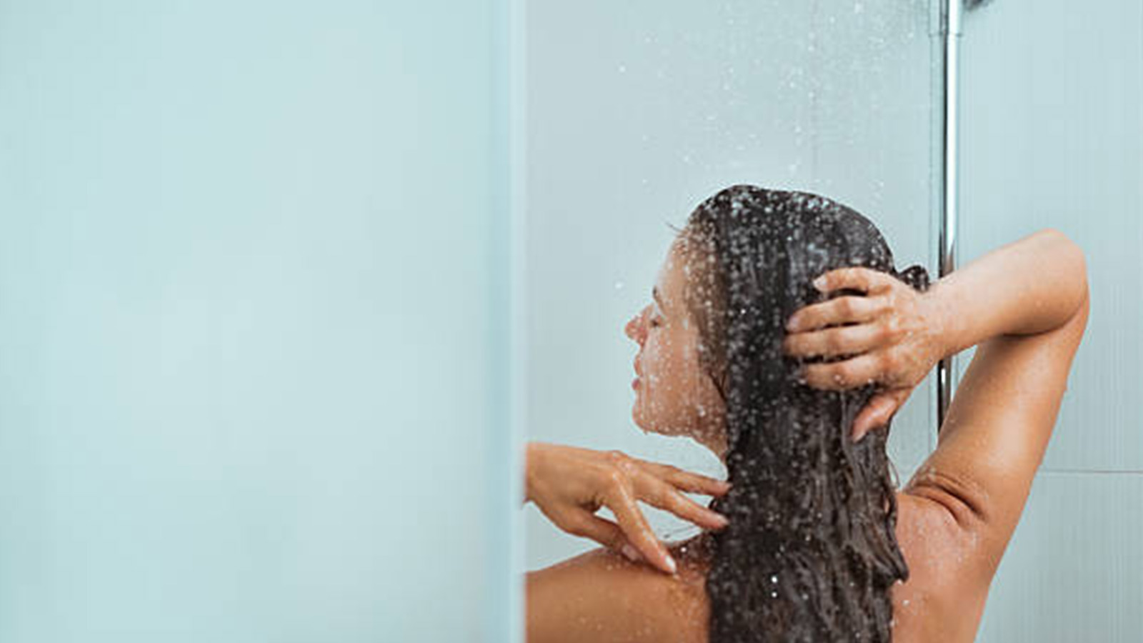 A woman is shown from behind, showering as she runs her hands through her long, dark hair. 