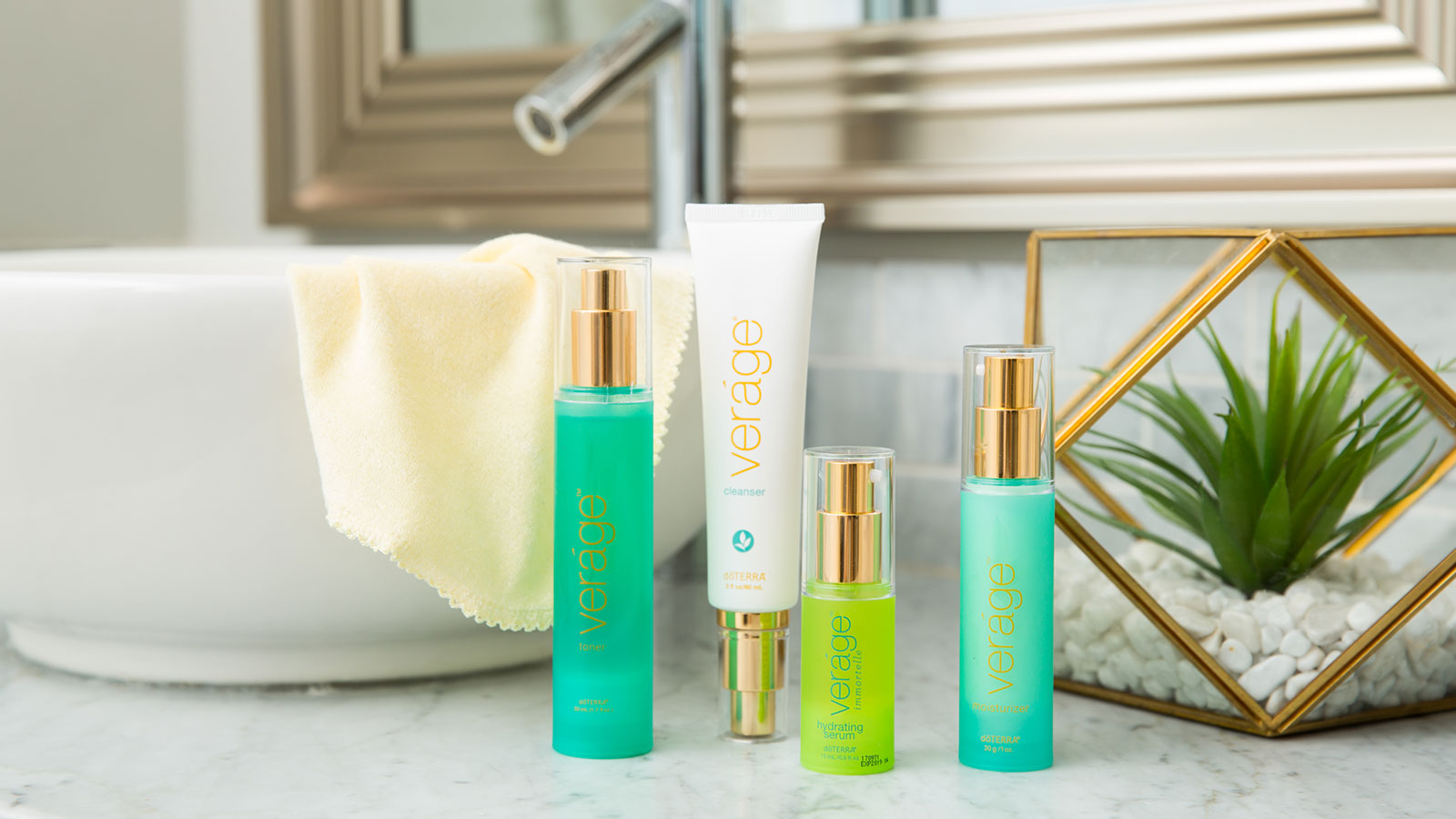 doTERRA Veráge Skin Care Collection sitting on a marble counter. Beside them is a green plant. Behind the collection is a large, bowl-shaped sink and a mirror.  