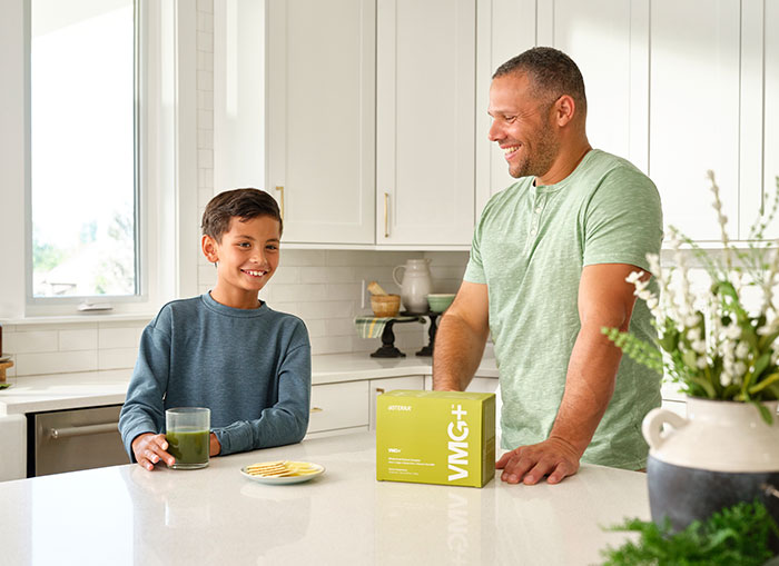 Father and son leaning against white kitchen countertop. VMG+ box sits by the father’s hand. The son has his hand around a clear cup filled with VMG+. A plate of cheese and crackers sits between the two of them. 