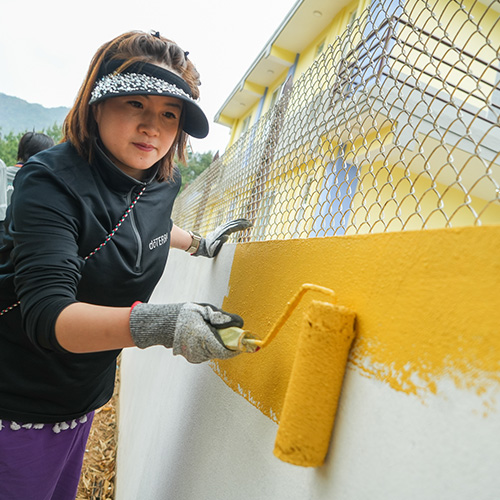 Woman outside, leaning her left hand for support on white cement wall, while using her right hand to paint the wall yellow with a roller brush. 