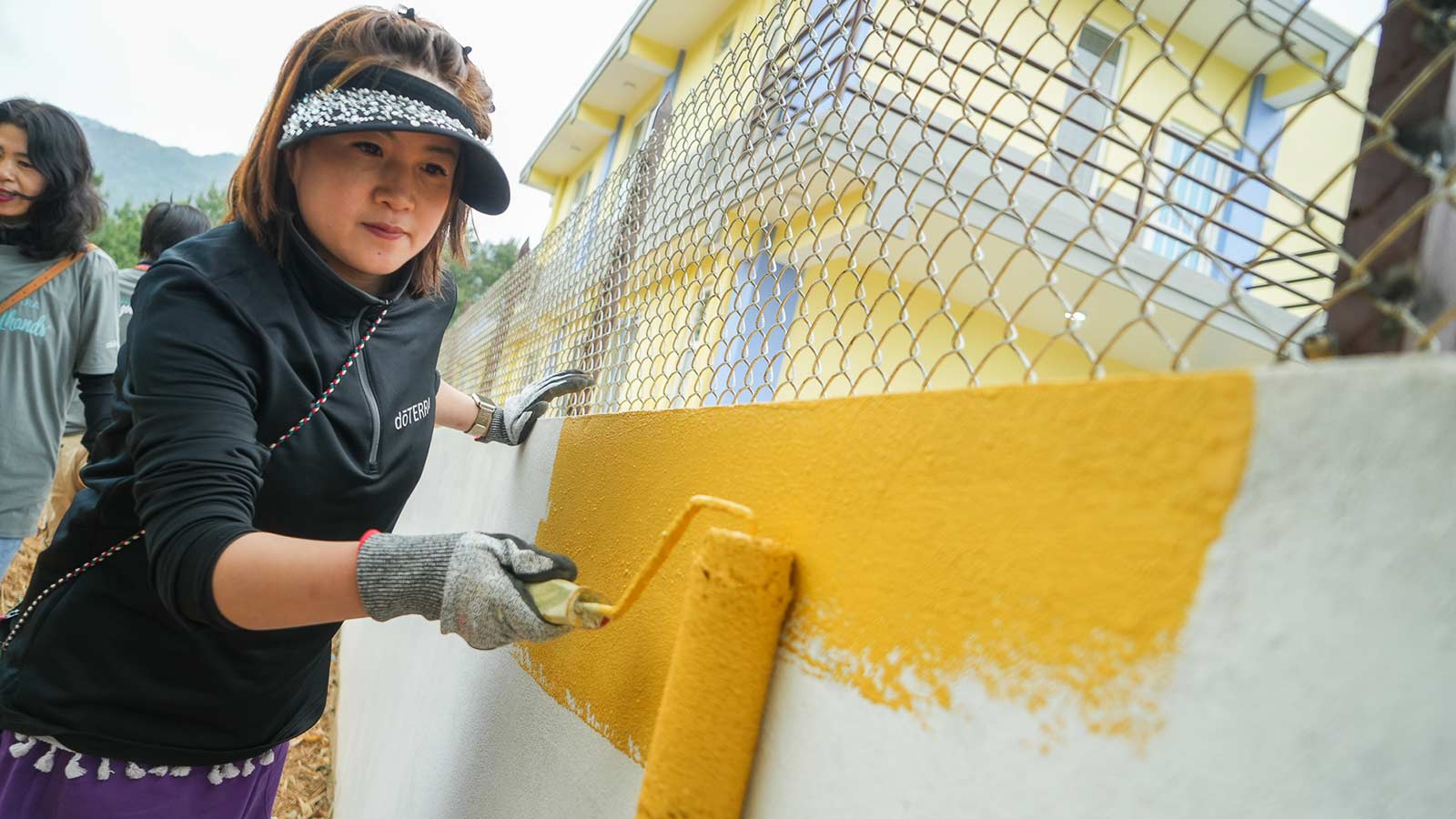 Woman outside, leaning her left hand for support on white cement wall, while using her right hand to paint the wall yellow with a roller brush. 