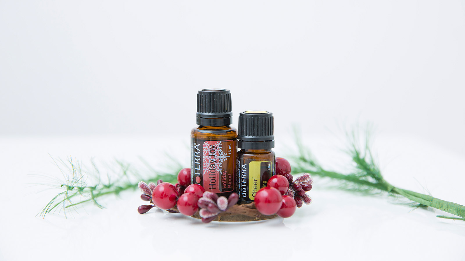 doTERRA Holiday Joy essential oil blend and doTERRA Cheer essential oil sit on a white surface surrounded by twigs and red cherry blossoms. A green branch is placed behind the essential oils.