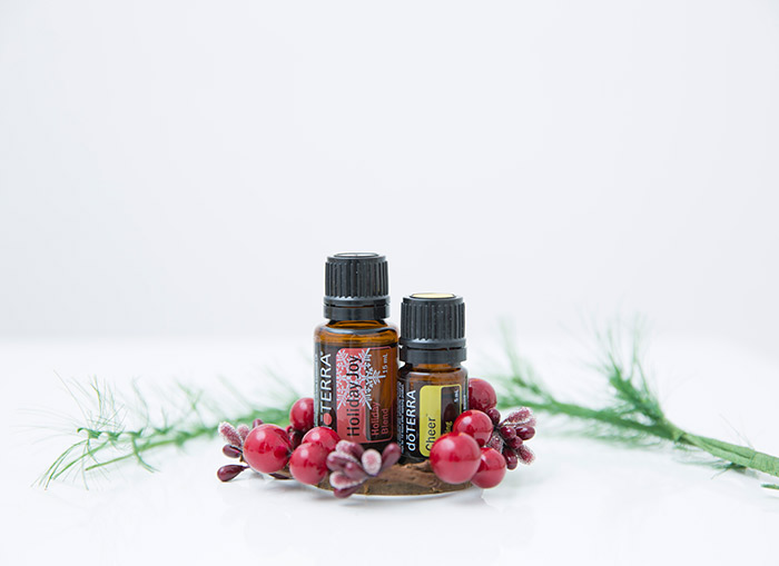 doTERRA Holiday Joy essential oil blend and doTERRA Cheer essential oil sit on a white surface surrounded by twigs and red cherry blossoms. A green branch is placed behind the essential oils.