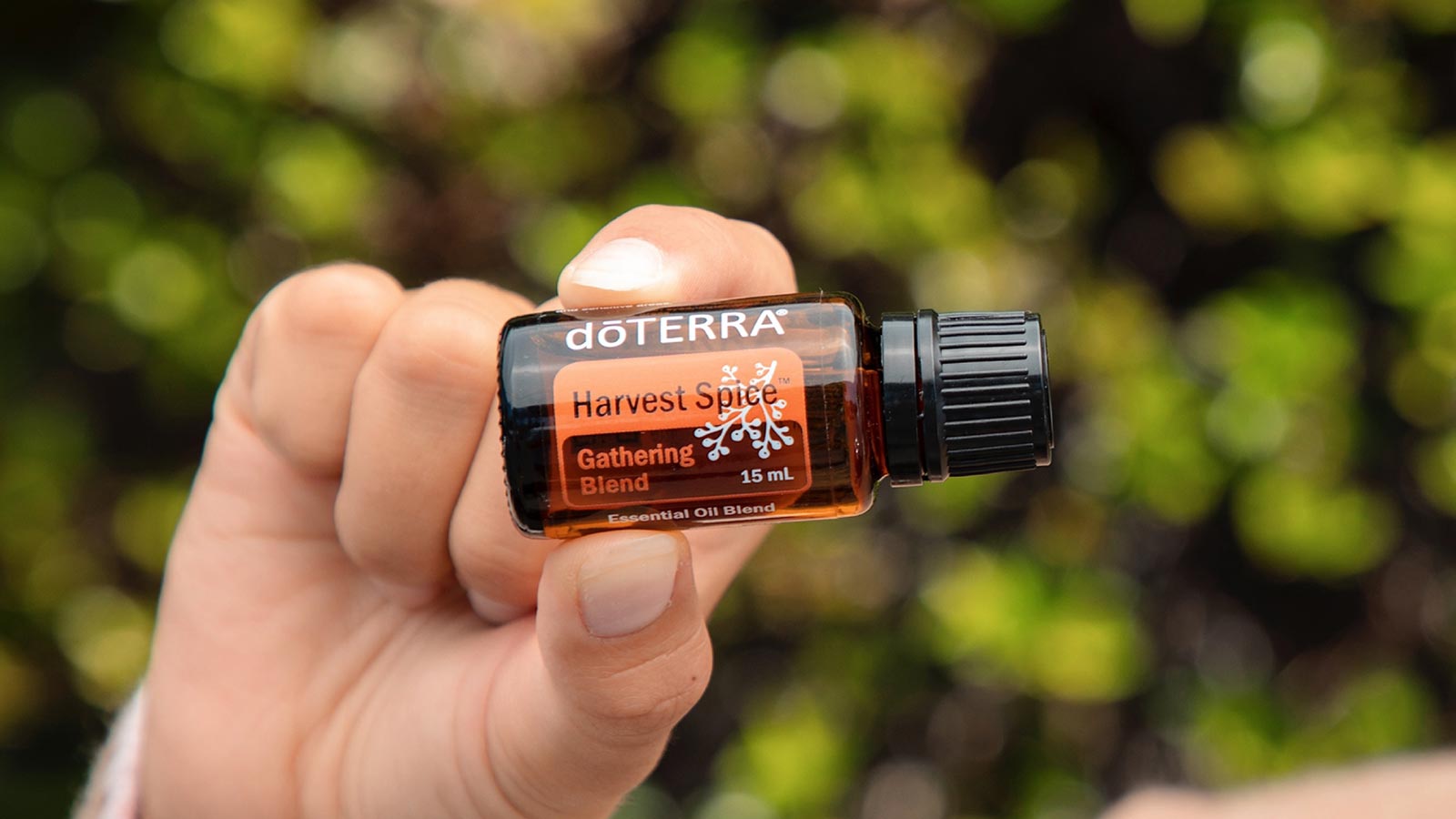 Someone holding doTERRA’s Harvest Spice essential oil blend with two fingers. Blurred outdoor scenery in the background.  