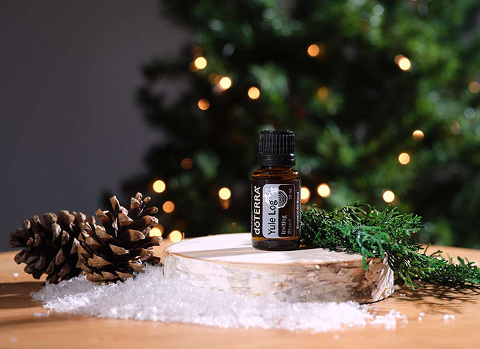 Yule Log blend seated on a small, round piece of wood, placed on a brown, round table. Pinecones, fake snow, and twigs surrounding the display. A lit Christmas tree is in the background. 