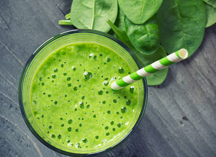 An overhead shot of a green drink in a glass cup with a green and white striped straw in the cup. The cup is on a gray surface with spinach sitting to the side. 