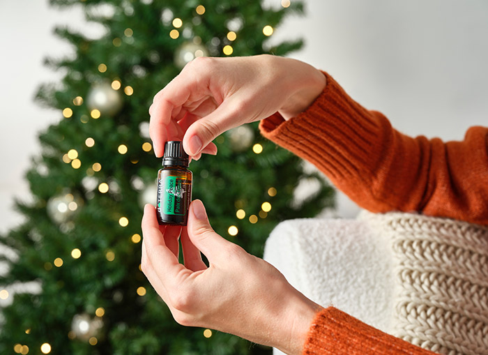 Woman holding doTERRA Holiday Peace essential oil blend, while resting her elbow on the armrest of a white couch. A lit Christmas tree in the background. 