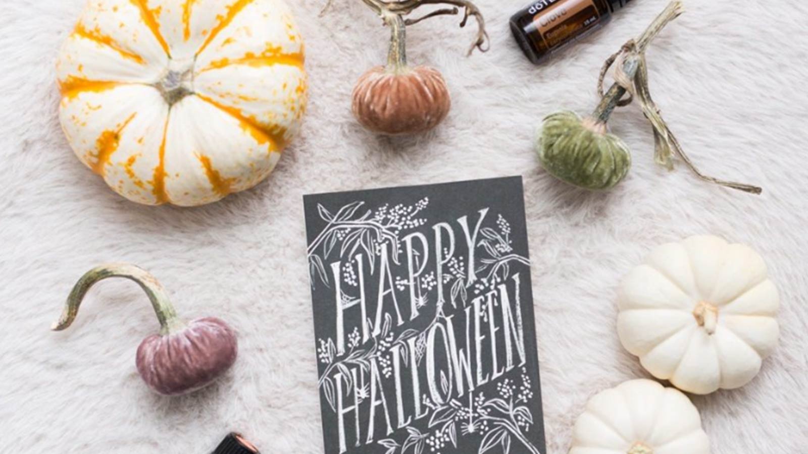 A black Happy Halloween card placed on a white, soft surface, surrounded by pumpkins and essential oil bottles. 