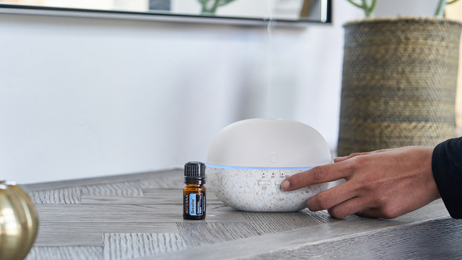 doTERRA Breathe sitting on a gray, rectangular table. The doTERRA Pebble Diffuser is sitting beside it. A woman is pressing the on button as the diffuser begins to diffuse.