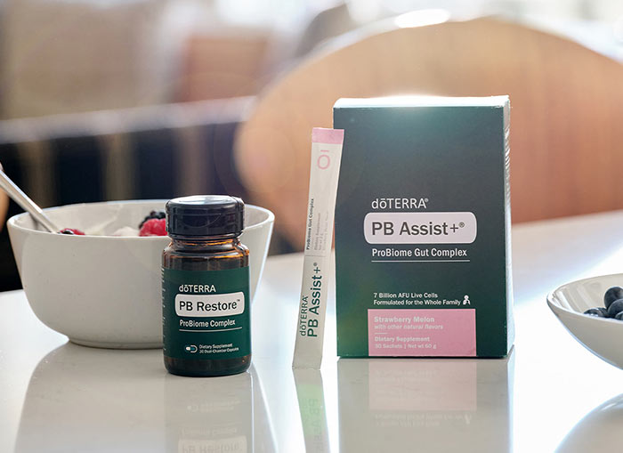 doTERRA PB Restore and PB Assist+ ProBiome Gut Complex sitting on a white counter with white bowls filled with berries. A clear glass cup of water is off the side. Wooden chairs are in the background.