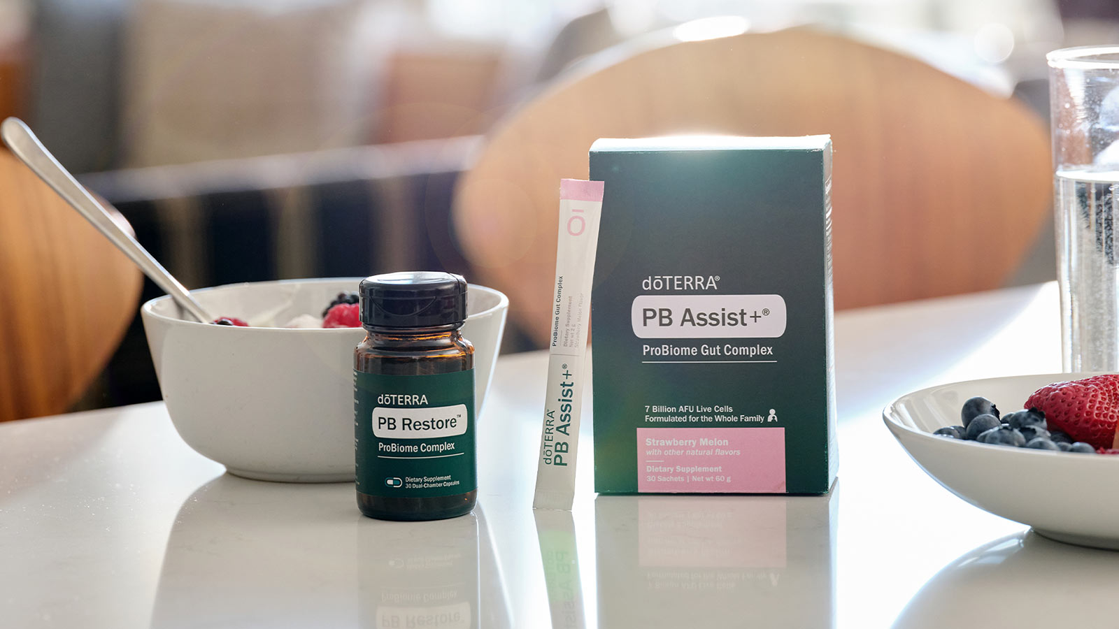 doTERRA PB Restore and PB Assist+ ProBiome Gut Complex sitting on a white counter with white bowls filled with berries. A clear glass cup of water is off the side. Wooden chairs are in the background.