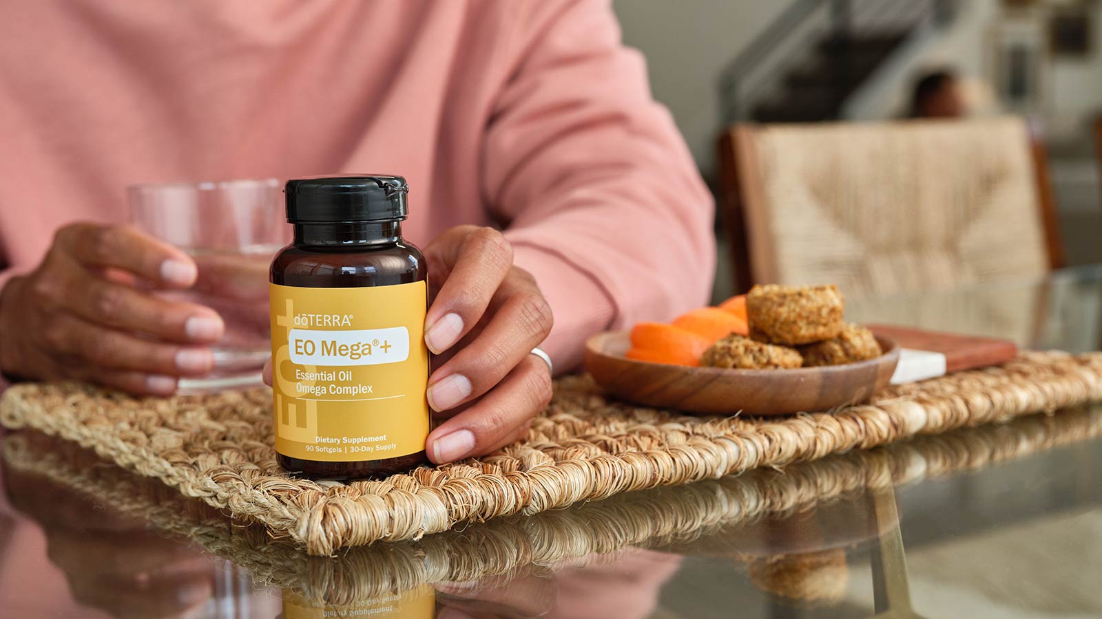 Person sitting at a kitchen table, holding a cup of water in their right hand, with their left around the EO Mega+ Softgels bottle. A plate of food sitting on a woven brown placement to their left.
