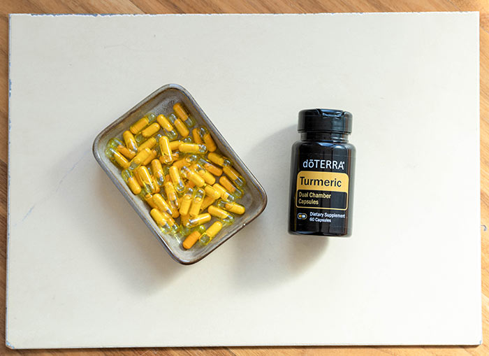 doTERRA Turmeric Dual Chamber Capsules and a tin full of yellow capsules, lying on a white mat that’s been placed on a wooden surface. 