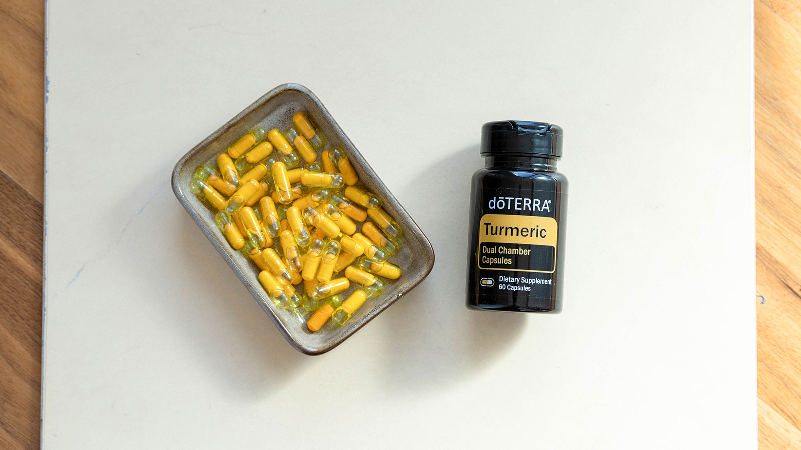 doTERRA Turmeric Dual Chamber Capsules and a tin full of yellow capsules, lying on a white mat that’s been placed on a wooden surface. 