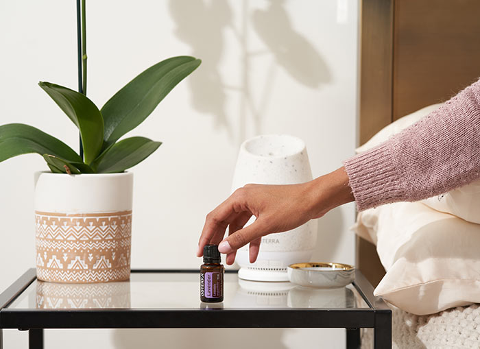 Woman, sitting on a bed, reaching for doTERRA Lavender essential oil on nightstand table. The doTERRA Roam Diffuser and a large green plant behind the oil.  
