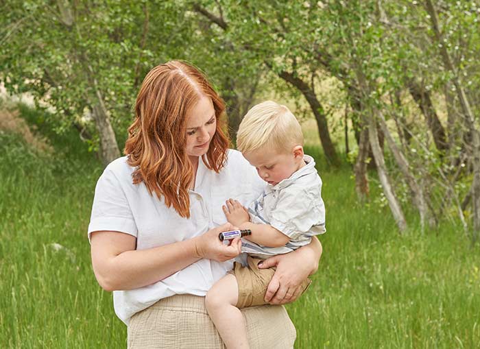 A woman stands outside in green grass, holding her son, while applying doTERRA Calmer Touch to her son’s arm. 