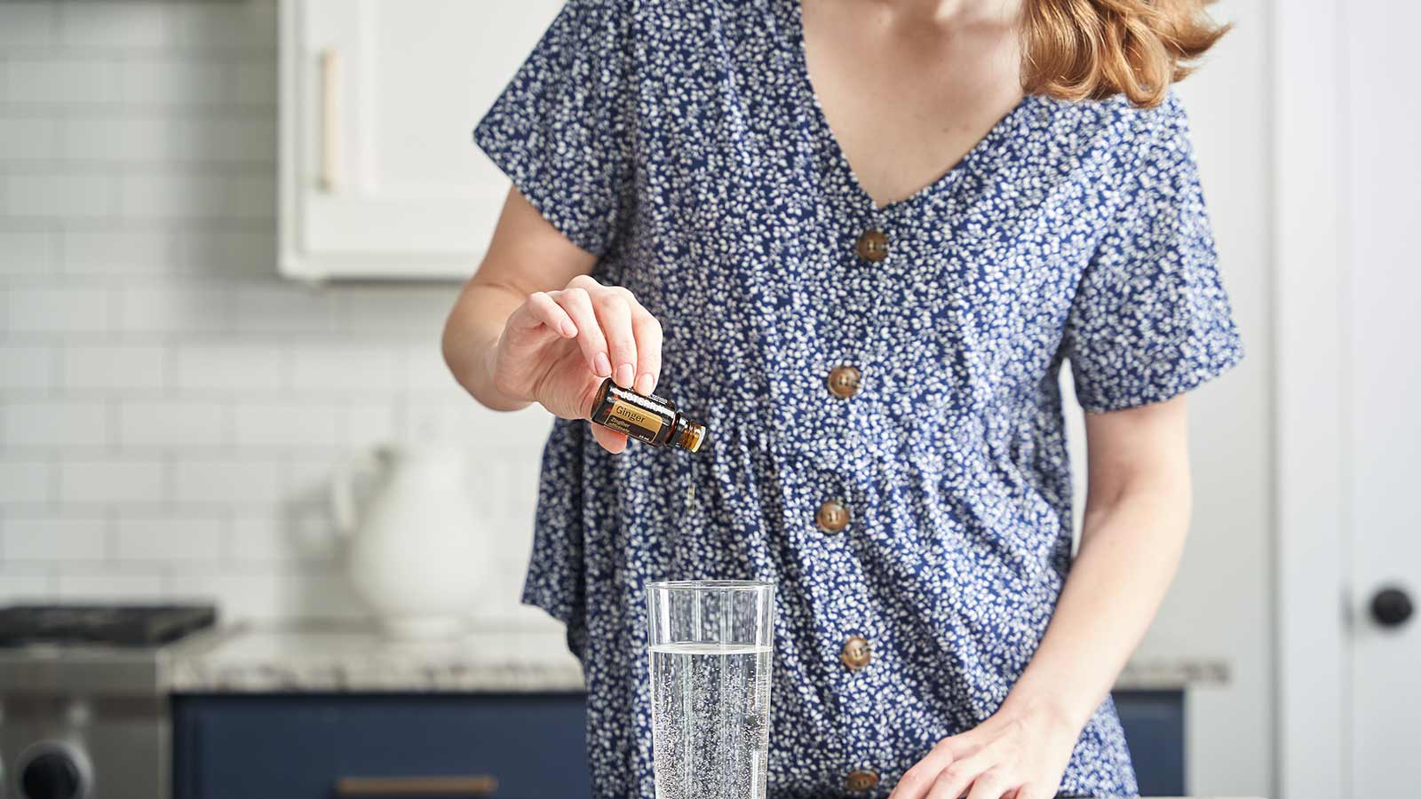 A woman standing next to a marble kitchen counter, her hand rested on the counter, and her other hand holding a bottle of doTERRA Ginger essential oil, pouring it into a glass of water.  
