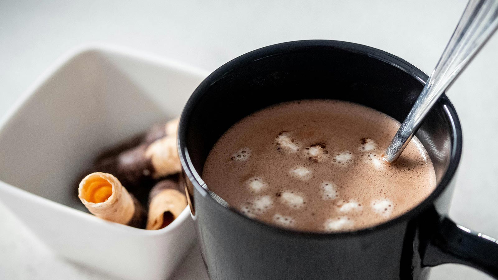 A black cup of hot chocolate, filled with marshmallows, sits on a counter. A square bowl of puff and chocolate pastries sits to the side. 