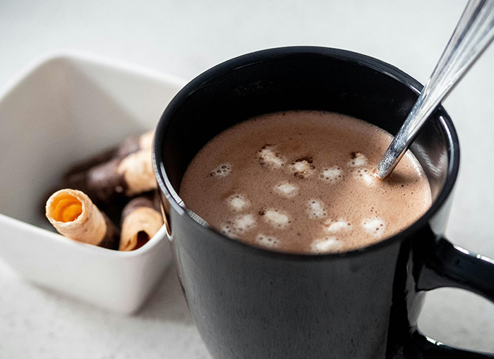 A black cup of hot chocolate, filled with marshmallows, sits on a counter. A square bowl of puff and chocolate pastries sits to the side. 