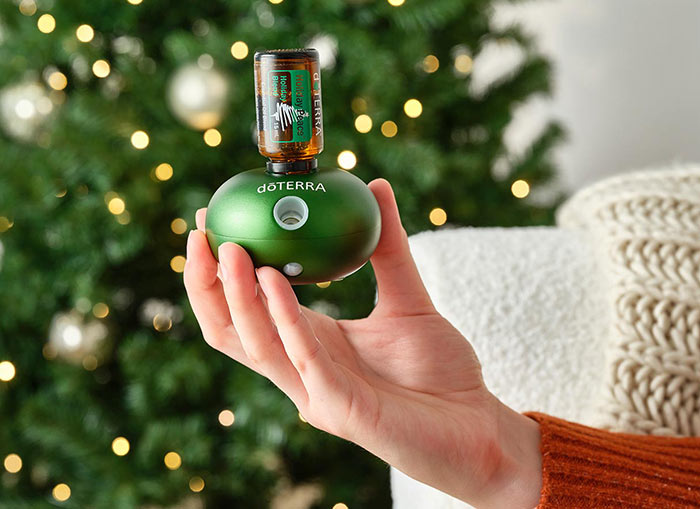 Someone holding Bubble Emerald Diffuser with Holiday Peace plugged in. A white couch with a cream blanket draped over and a lit Christmas tree in the background.