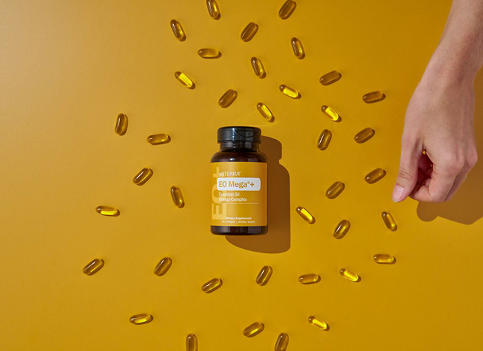 doTERRA EO Mega+ lying on a yellow surface with several of the capsules scattered about. A hand stretched out to pick up one of the pills. 