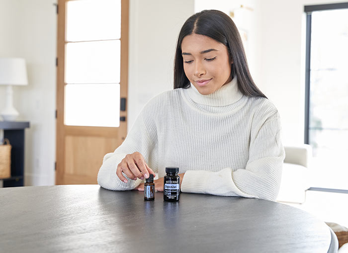 A woman is sitting at a round table while gently touching doTERRA DigestZen essential oil blend and DigestZen Softgels sit beside the bottle.  