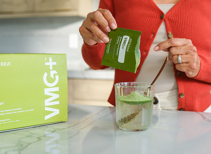 Women pouring VMG+™ Whole-Food Nutrient Complex into a cup of water. Package of VMG+™ sitting beside cup on marble counter.
