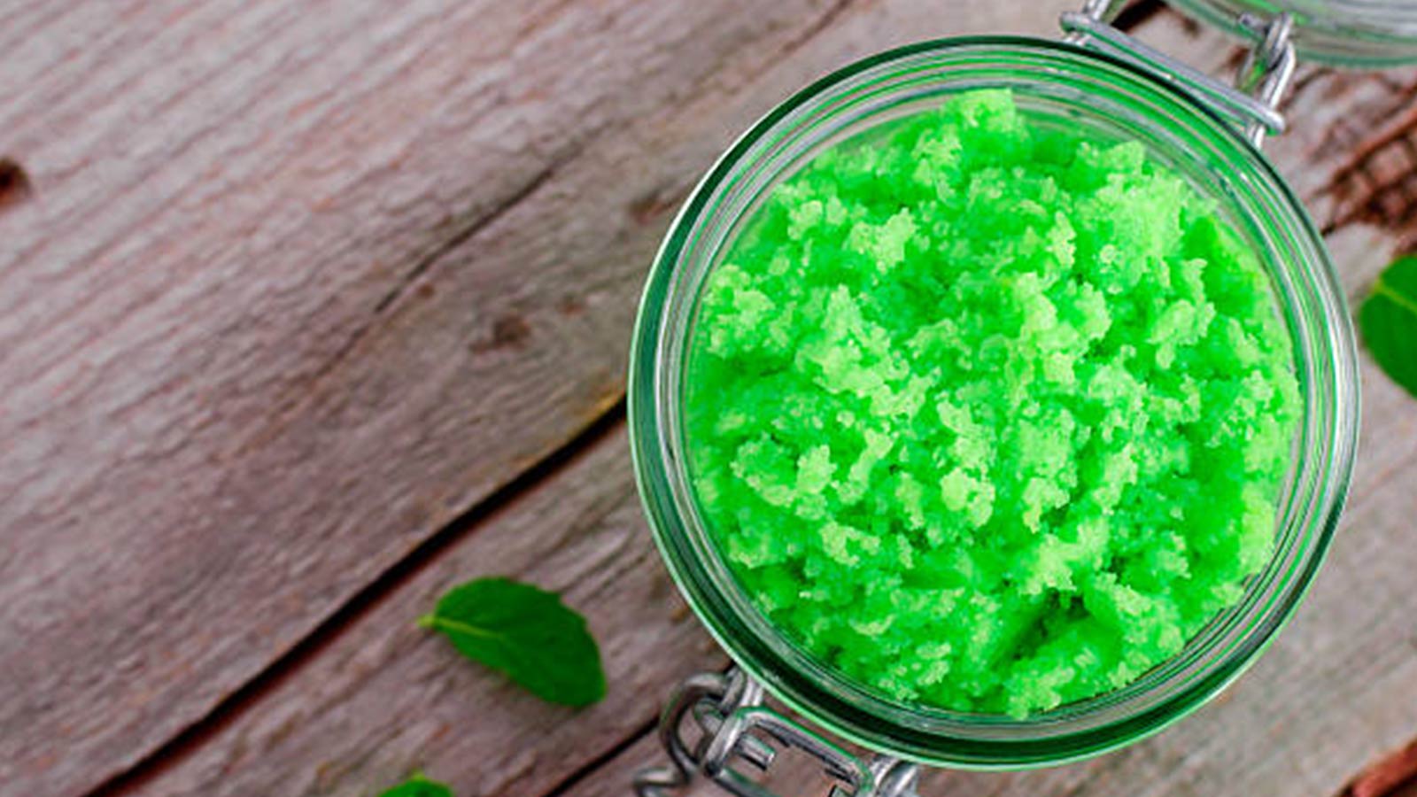 An overhead shot of green sugar scrub in a glass, round jar. The jar is sitting on a wooden surface. 
