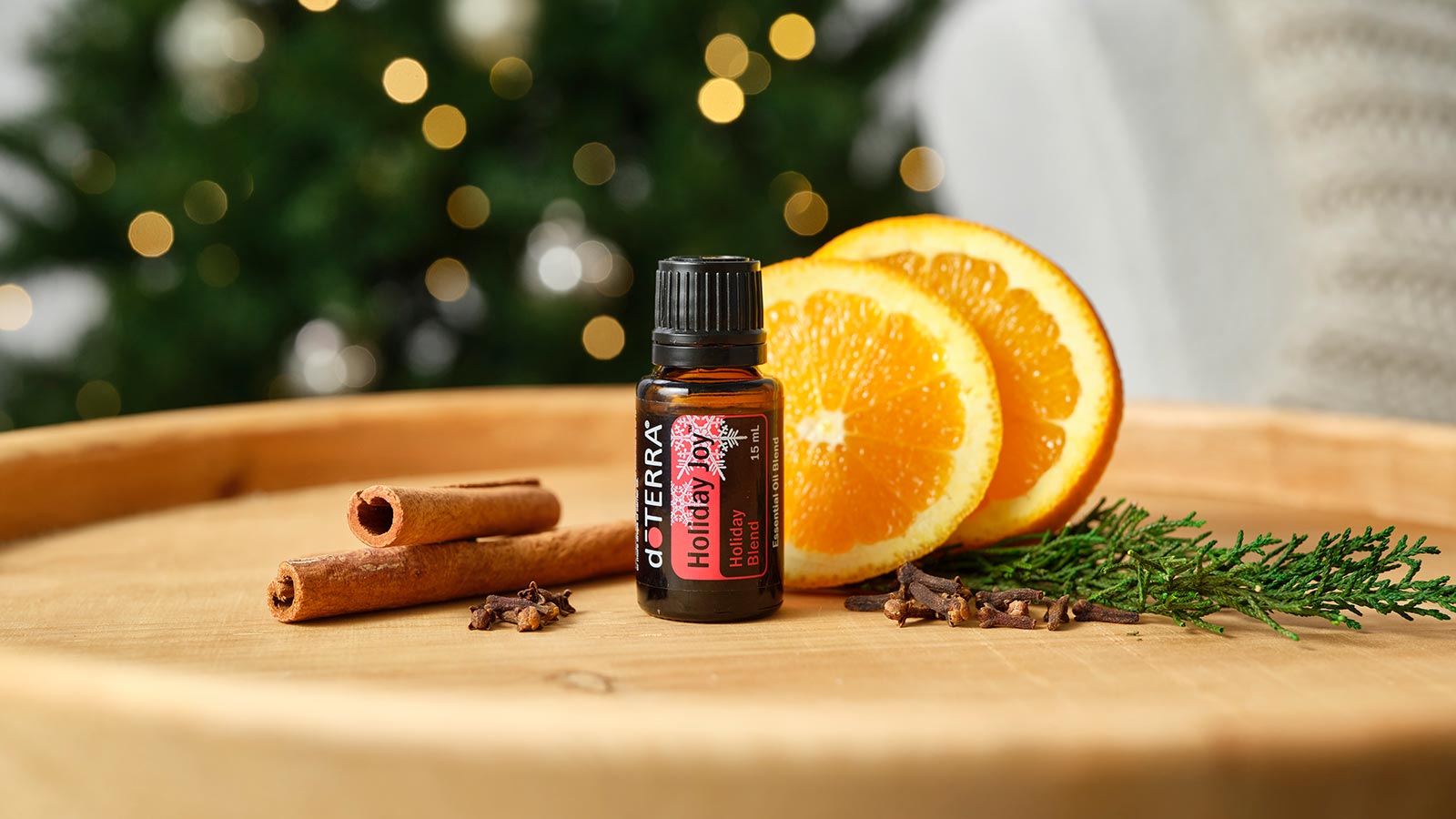 doTERRA Holiday Joy essential oil blend is seated on a round, wooden table. Sliced oranges, cinnamon sticks and buds, and a pine tree branch decorate the area. 