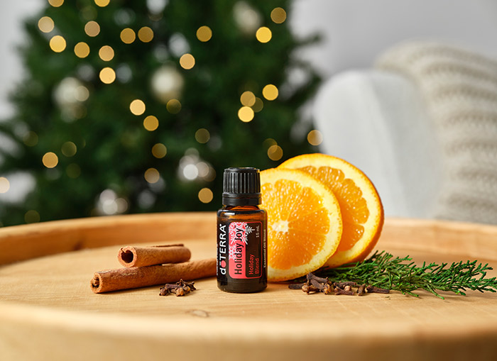 doTERRA Holiday Joy essential oil blend is seated on a round, wooden table. Sliced oranges, cinnamon sticks and buds, and a pine tree branch decorate the area. 