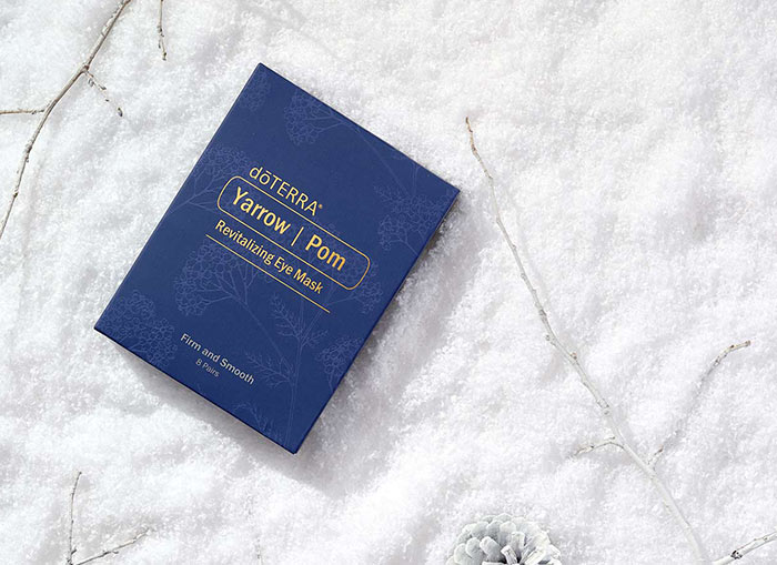 Yarrow Pom Eye Mask laying on a soft, snow-like surface surrounded by white twigs and pinecones.