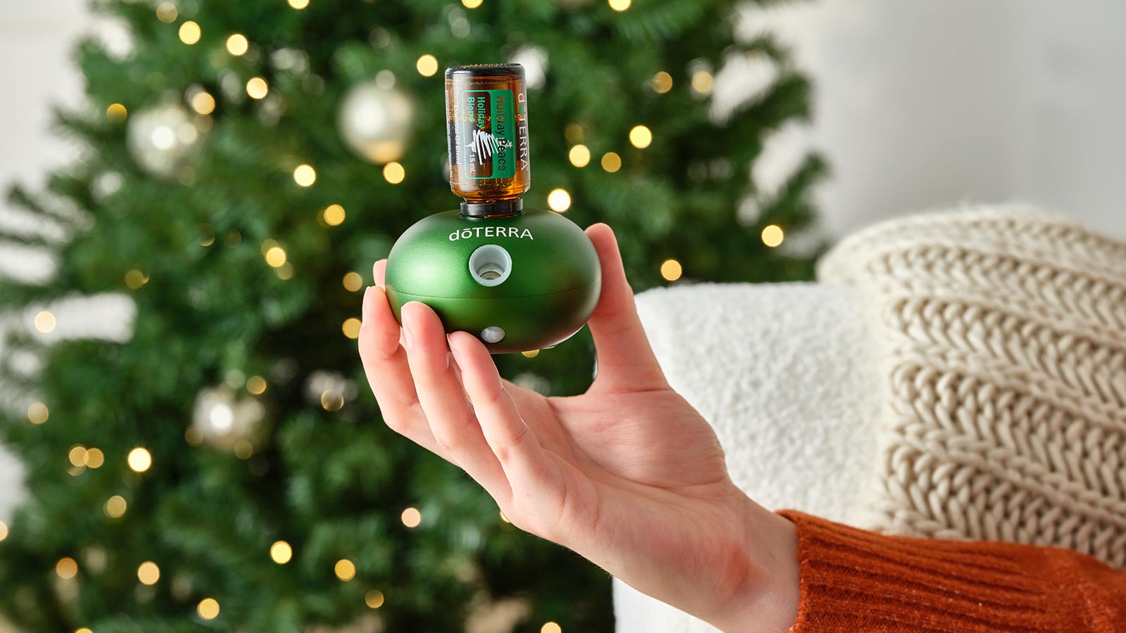 Someone holding Bubble Emerald Diffuser with Holiday Peace plugged in. A white couch with a cream blanket draped over and a lit Christmas tree in the background.