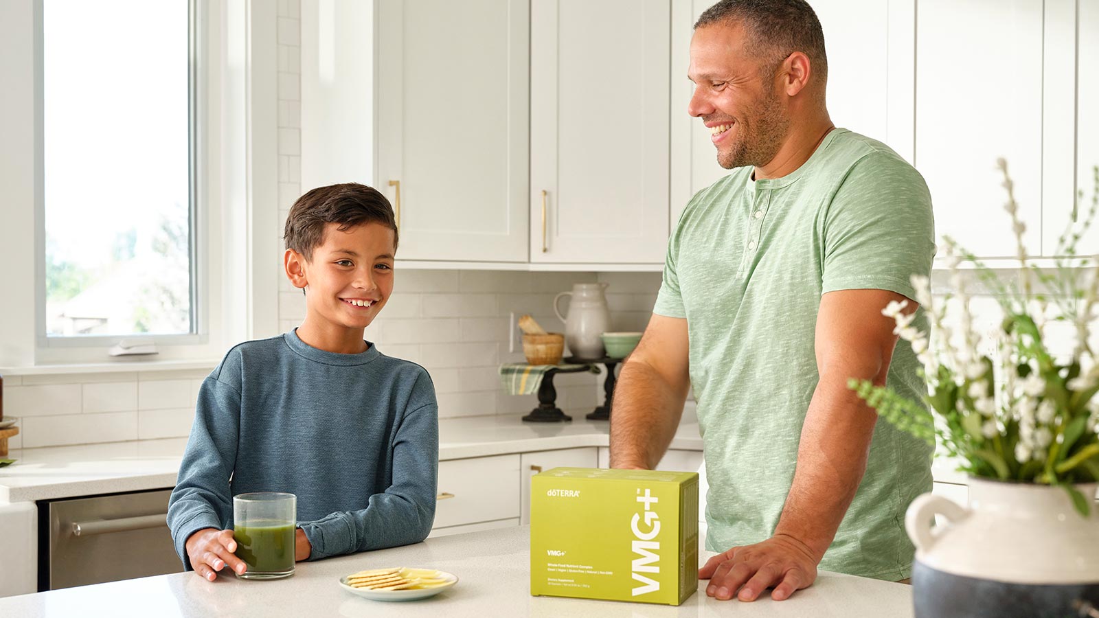 Father and son leaning against white kitchen countertop. VMG+ box sits by the father’s hand. The son has his hand around a clear cup filled with VMG+. A plate of cheese and crackers sits between the two of them. 