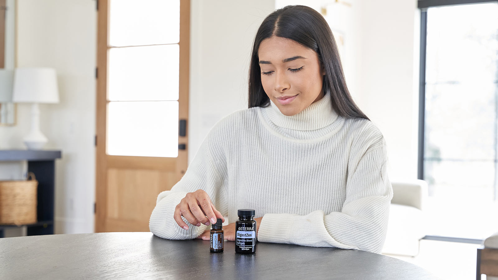 A woman is sitting at a round table while gently touching doTERRA DigestZen essential oil blend and DigestZen Softgels sit beside the bottle.  