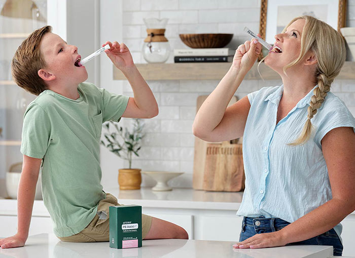 Son and mother are pouring PB Assist+ ProBiome Gut Complex sachets into their mouths in a primarily white kitchen. The son sits on the counter, with the mother standing beside him.