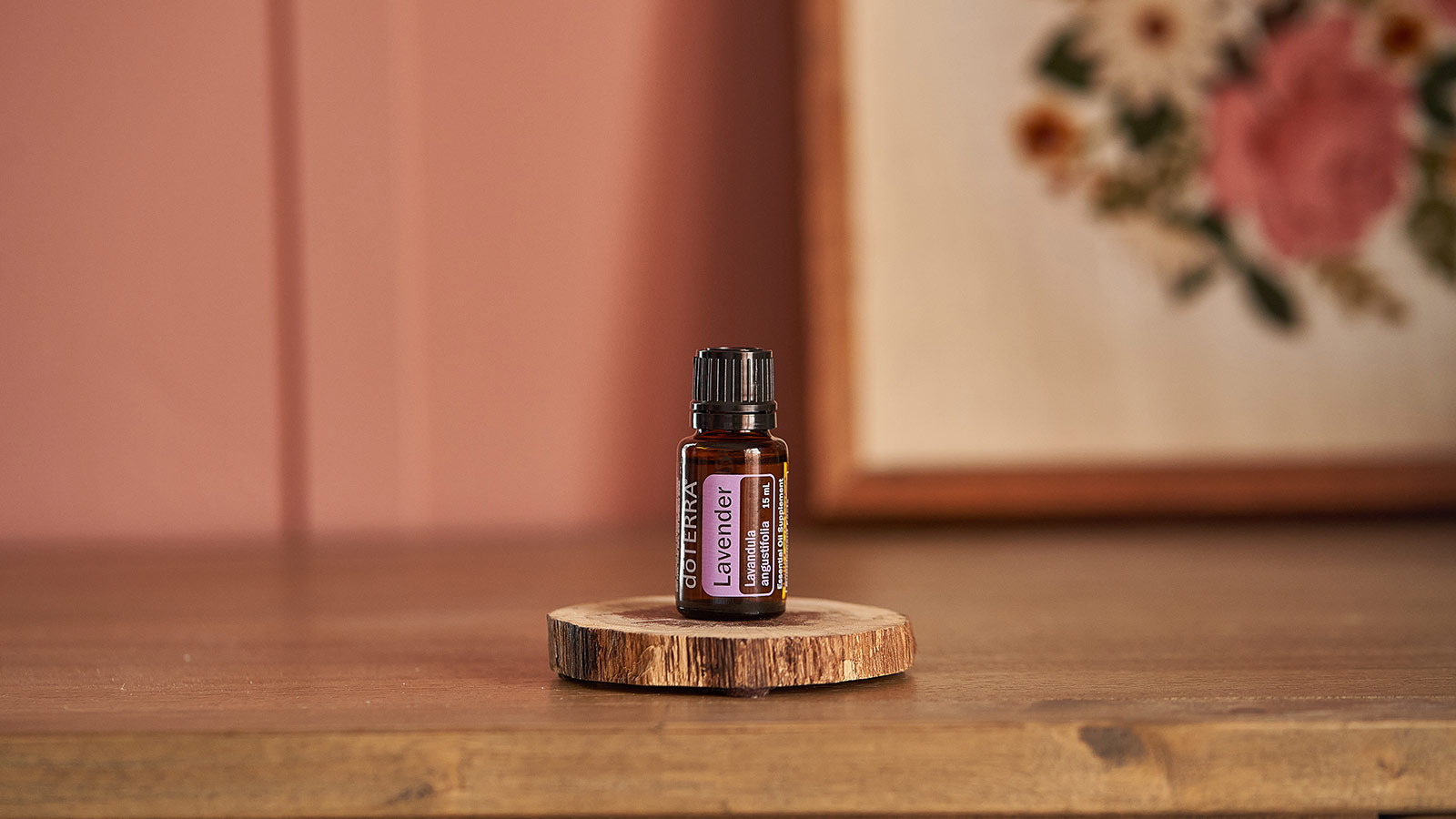 doTERRA Lavender essential oil seated on thin bark platform on a wooden table. A pink wall and floral needlework framed in the background. 