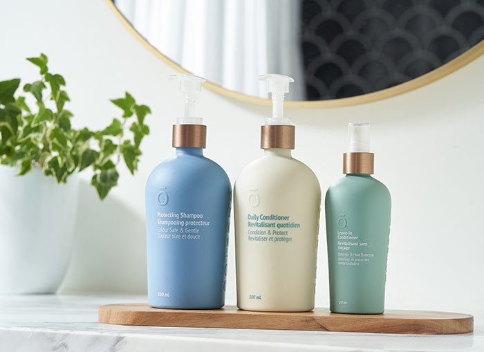 From left to right sits doTERRA Protecting Shampoo, Daily Conditioner, Leave-In Conditioner on a thin piece of shaped wood, placed on a marble counter. A green plant and mirror in the background. 