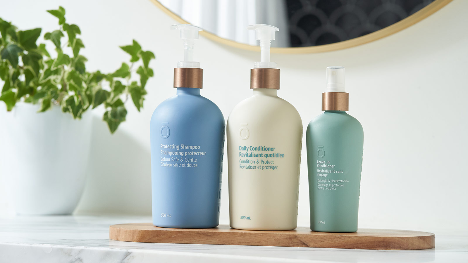 From left to right sits doTERRA Protecting Shampoo, Daily Conditioner, Leave-In Conditioner on a thin piece of shaped wood, placed on a marble counter. A green plant and mirror in the background. 