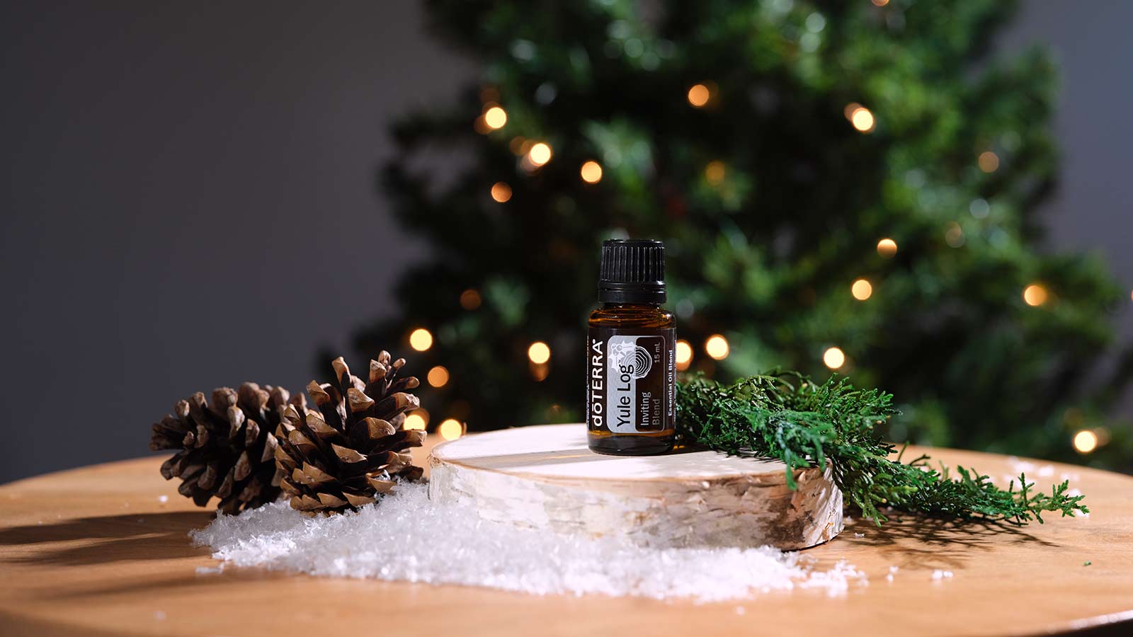Yule Log blend seated on a small, round piece of wood, placed on a brown, round table. Pinecones, fake snow, and twigs surrounding the display. A lit Christmas tree is in the background. 